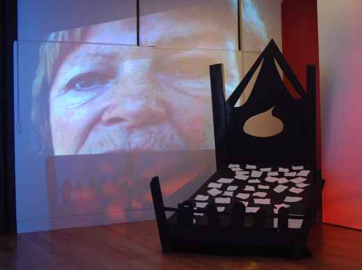 A giant gothic bed covered in scraps of paper with fears written on them; behind a projected image of a woman and a noose.